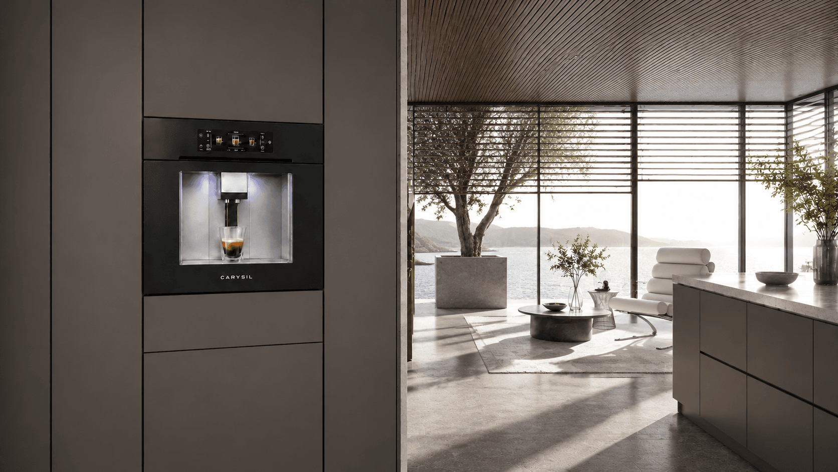 A Carysil built-in coffee machine pulling an espresso, set into a kitchen wall opening onto a lakeside living room